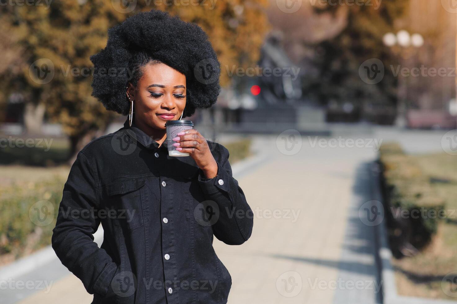glad mörk flådd kvinna njuter kaffe innehav till gå kopp återskapa i stad parkera, Lycklig trendig klädd afrikansk amerikan hipster flicka Sammanträde utomhus under solig dag dricka dryck foto