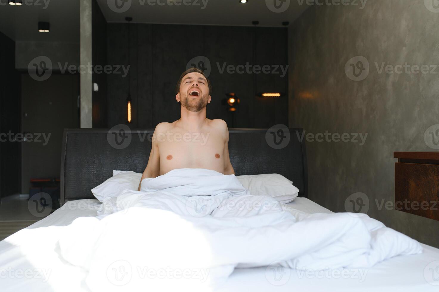 ung man vakna upp och stiga händer stretching på vit säng i de morgon- med solljus genom fönster foto