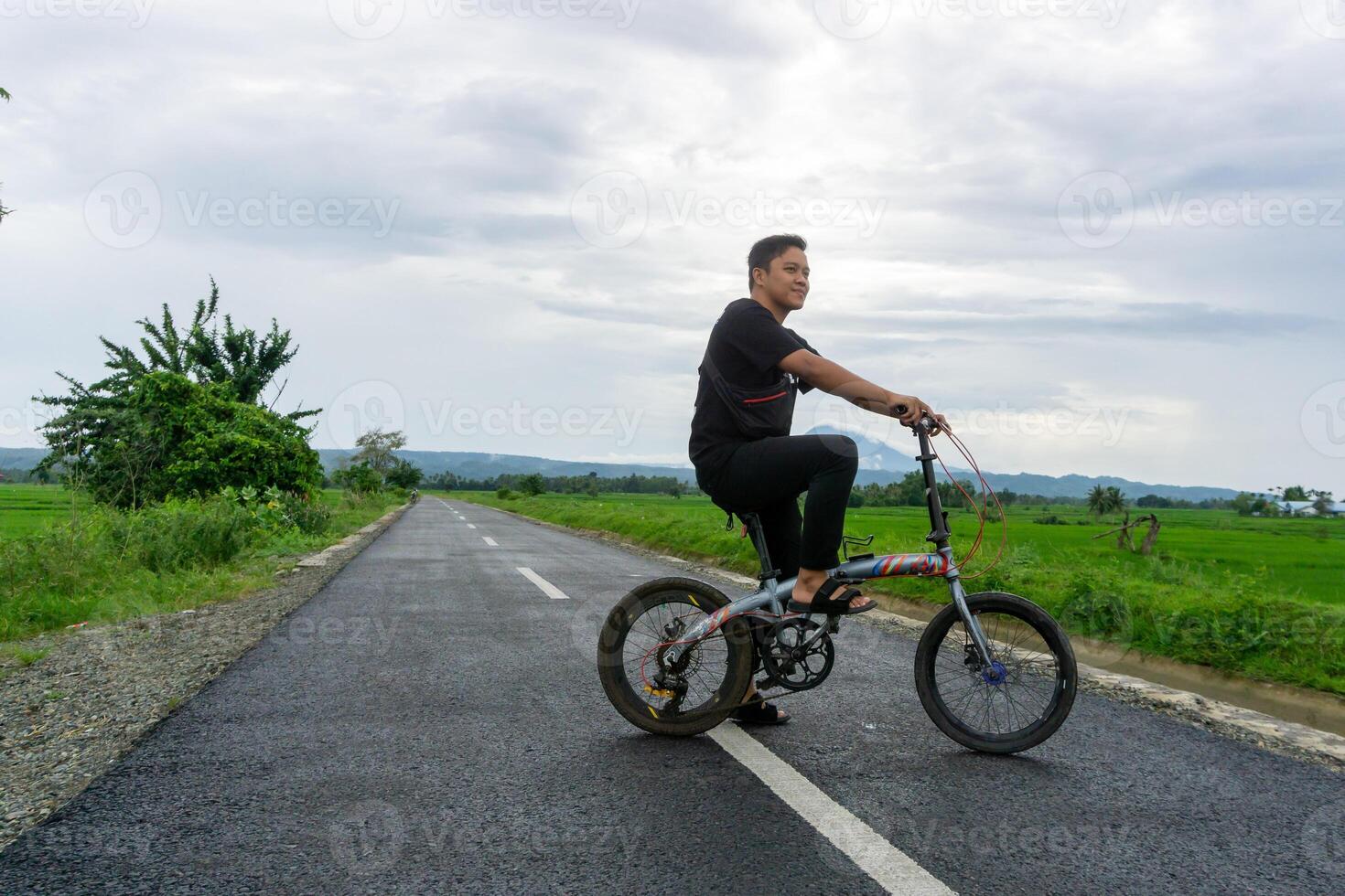 Lycklig asiatisk man ridning en cykel på de morgon- på de asfalt väg. cykling med berg och irländare ris fält se på de bakgrund. foto