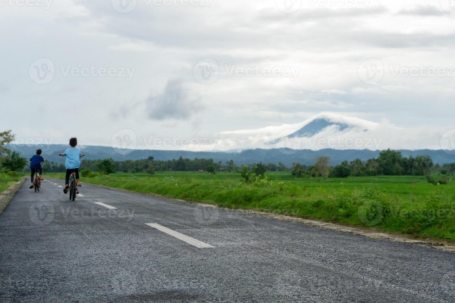 två asiatisk barn ridning cykel på de morgon- på de asfalt väg. två Pojkar cykling med berg och irländare ris fält se på de bakgrund. kopia Plats foto