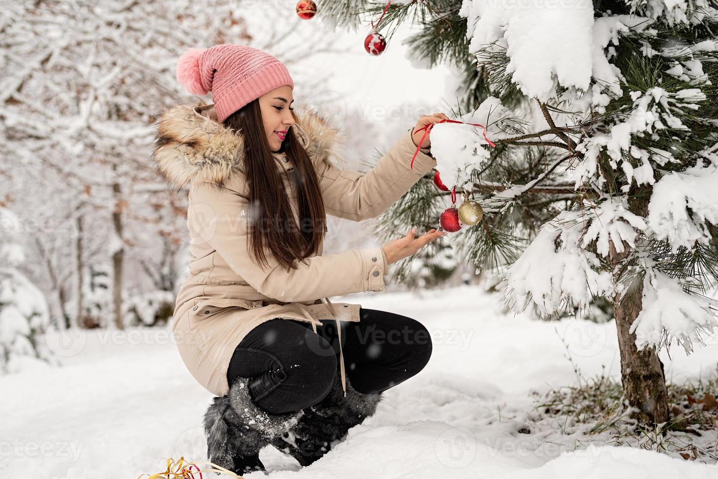 vacker kvinna i varma vinterkläder dekorerar julgran i en park i snöig dag foto