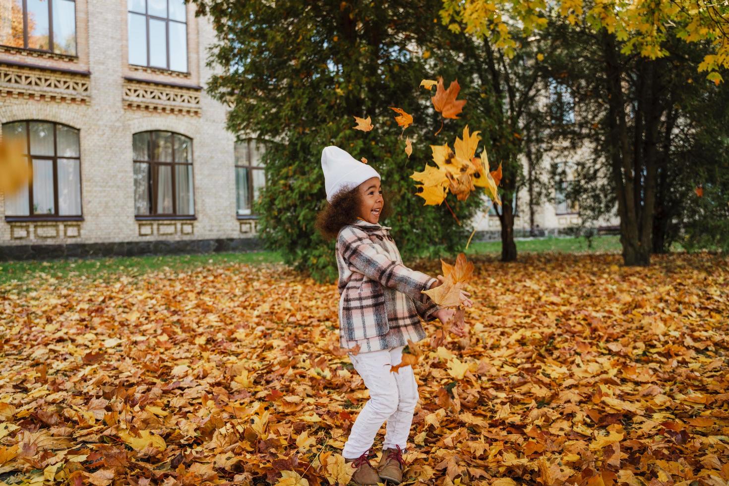 svart flicka bär kappa att göra kul med fallna löv i höst park foto