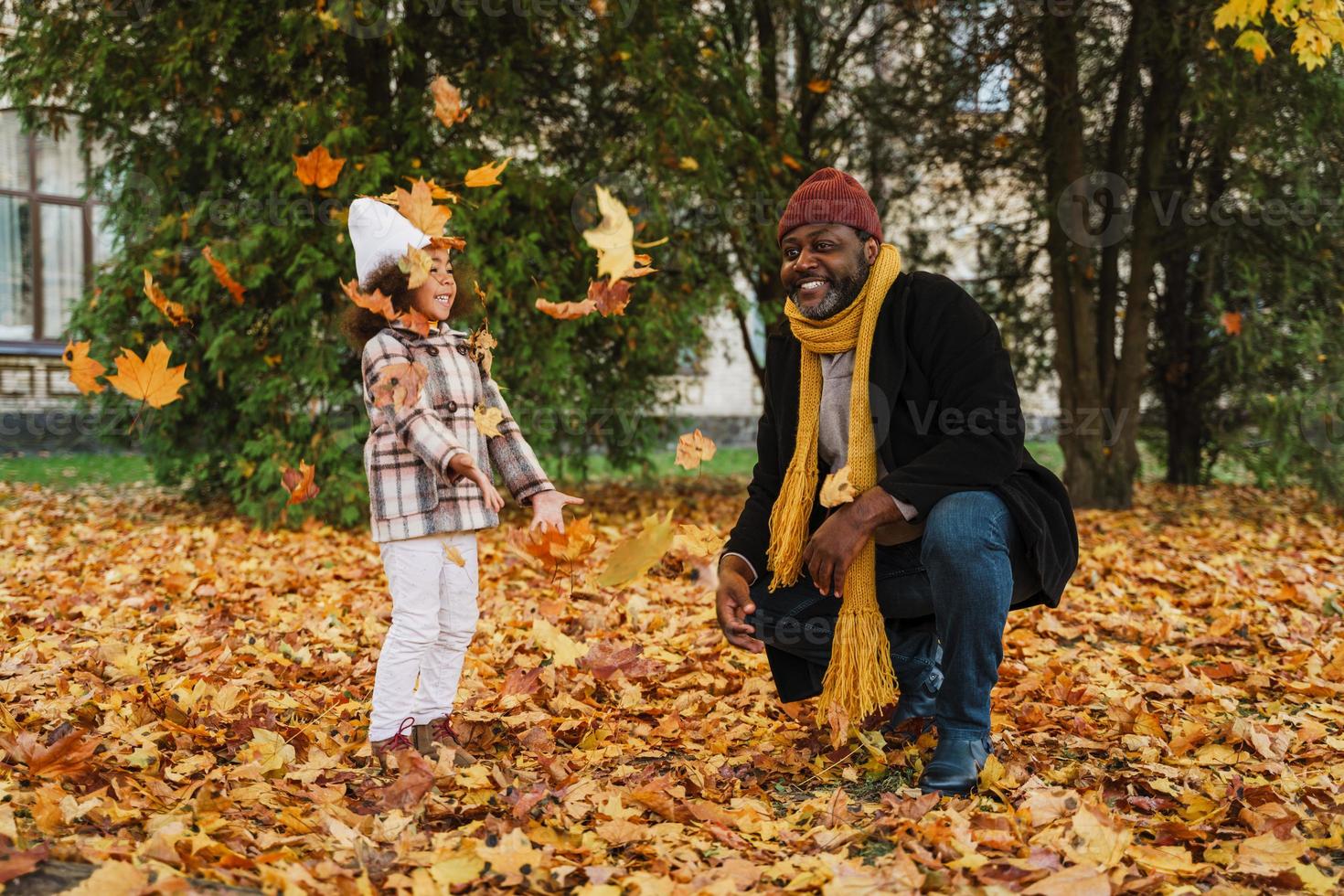 svart farfar och barnbarn gör kul med fallna löv i höst park foto