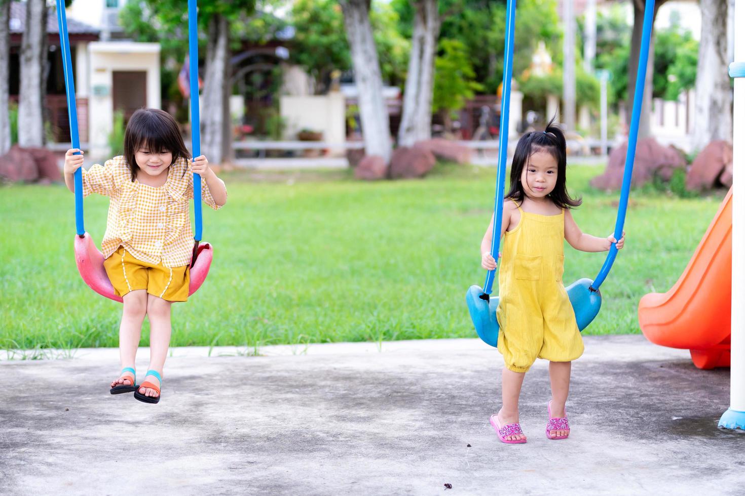 två asiatiska tjejer har kul när de svänger fram och tillbaka. söta barn bär gula kläder för att leka på lekplatsen på sommaren eller våren. barn i åldrarna 3 och 4 år. foto