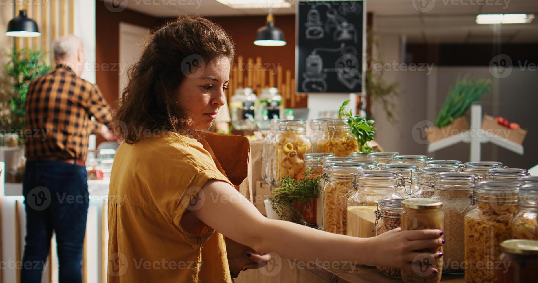 Lycklig kvinna i noll avfall mataffär använder sig av papper väska till inköp skafferi häftklamrar. vegan kund i lokal- grannskap matvaror affär håller på med handla för matlagning Ingredienser foto