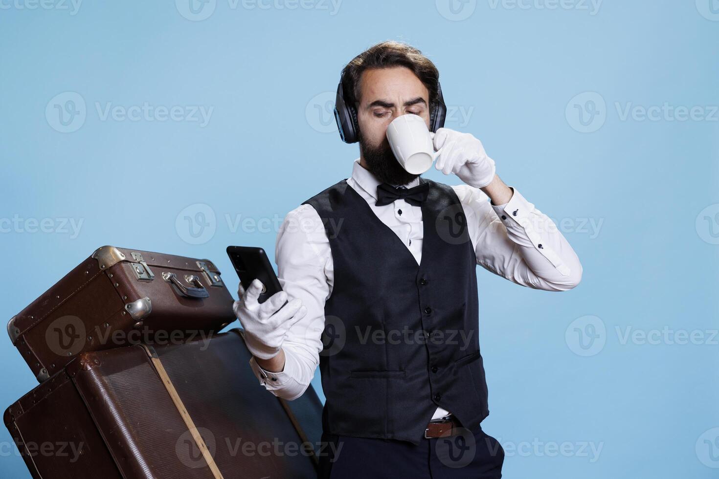 hotell porter drycker kaffe och sms, läsning meddelanden på smartphone och lyssnande till musik på hörlurar. portvakt bellboy med headsetet njuter dryck och bläddring webbsidor. foto