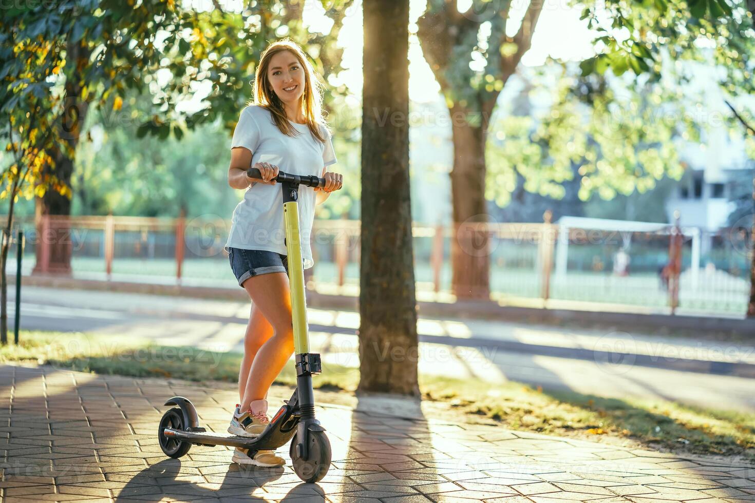 leende kvinna rider elektrisk skoter eller e-skoter i stad parkera på solnedgång. kvinna använder sig av elektrisk transport i urban parkera foto