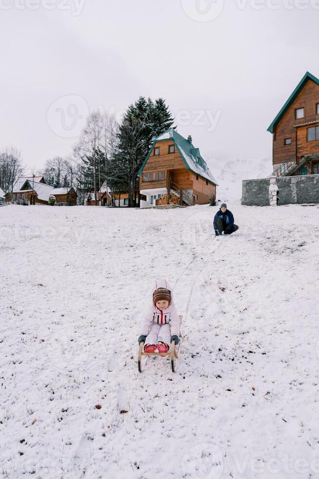 pappa knäböj på en kulle tittar på en liten flicka gå ner på en kälke foto