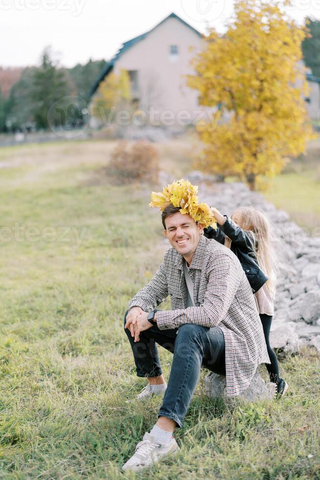 liten flicka sätter en krans av gul löv på de huvud av henne skrattande far Sammanträde på en sten i en clearing foto