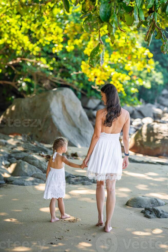 ung kvinna mor med en liten dotter i vit klänningar gående på havsstrand i de skugga av träd och handflatorna. hög kvalitet Foto