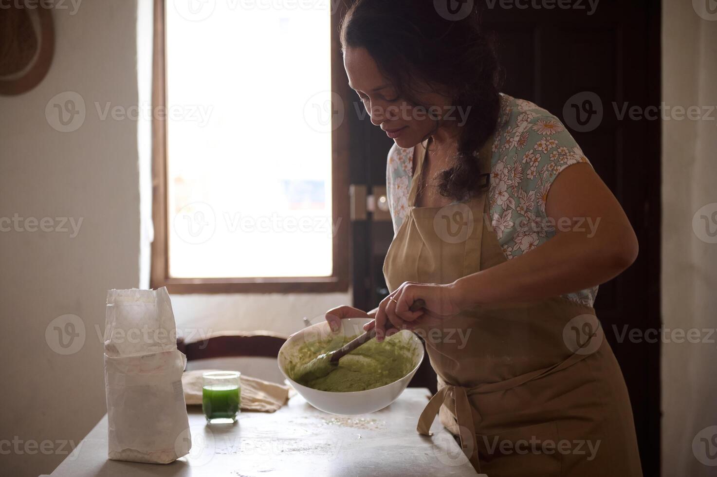 underbar kvinna kock, hemmafru i beige förkläde, stående förbi kök tabell, knådning deg med trä- sked, framställning traditionell ravioli enligt till traditionell italiensk recept, i lantlig miljö foto