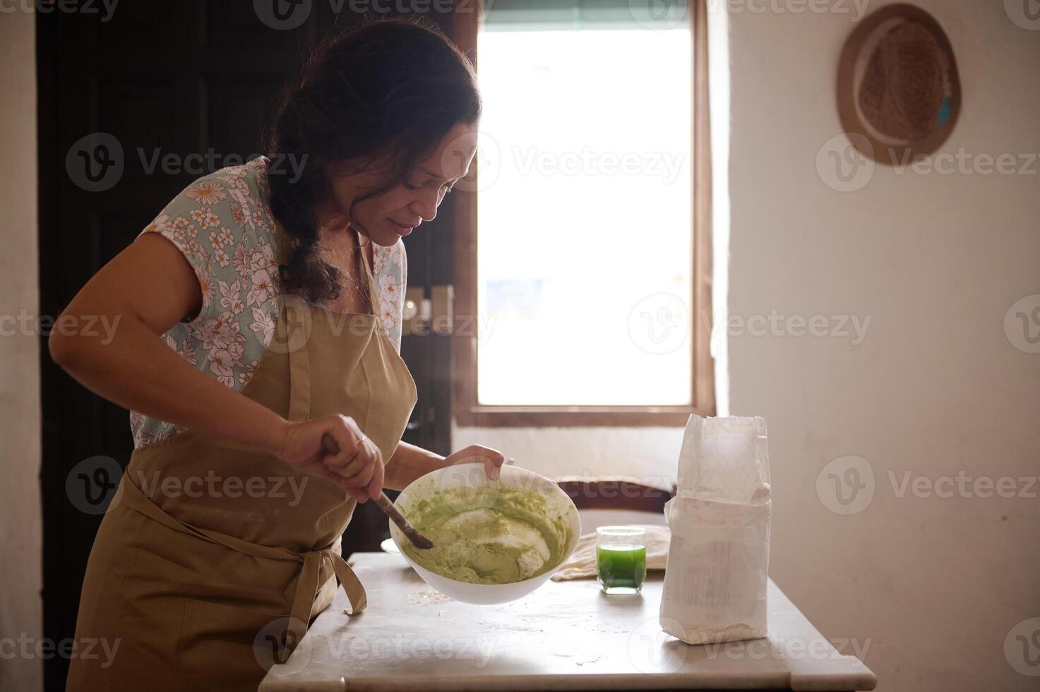 äkta Söt kvinna i beige förkläde, stående förbi kök tabell, knådning deg med trä- sked, framställning traditionell ukrainska maträtt varenyky enligt till traditionell recept, i de rustik kök foto