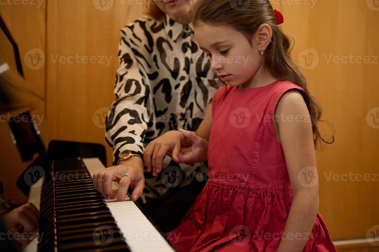 självsäker porträtt av en skön caucasian liten barn flicka i elegant röd klänning, inlärning spelar på pianoforte. kvinna pianist lärare förklara lektion, Sammanträde tillsammans på stor piano foto