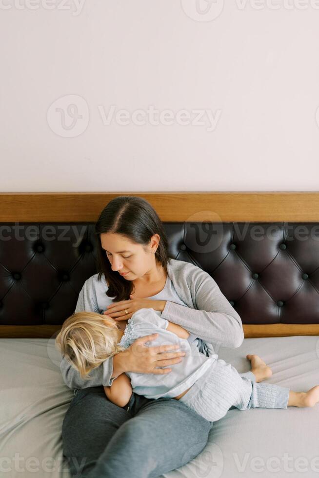 mamma ammar en liten flicka kramas henne i henne vapen medan Sammanträde på de säng foto