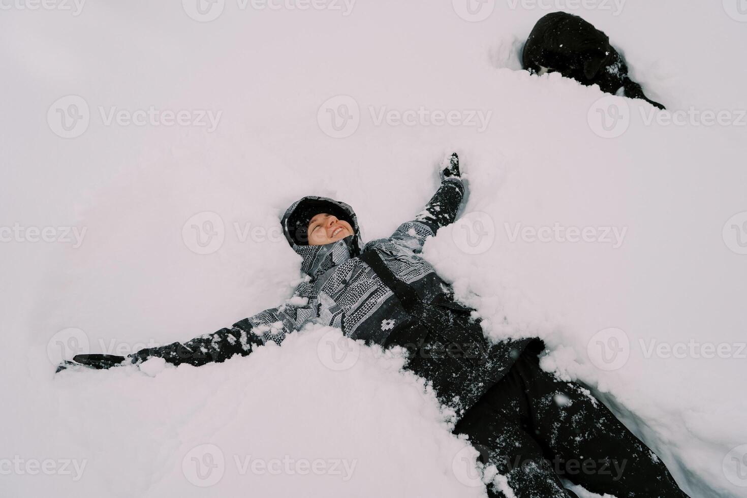 ung leende kvinna i en åka skidor kostym lögner i de snö med henne vapen utsträckt nära en svart hund foto