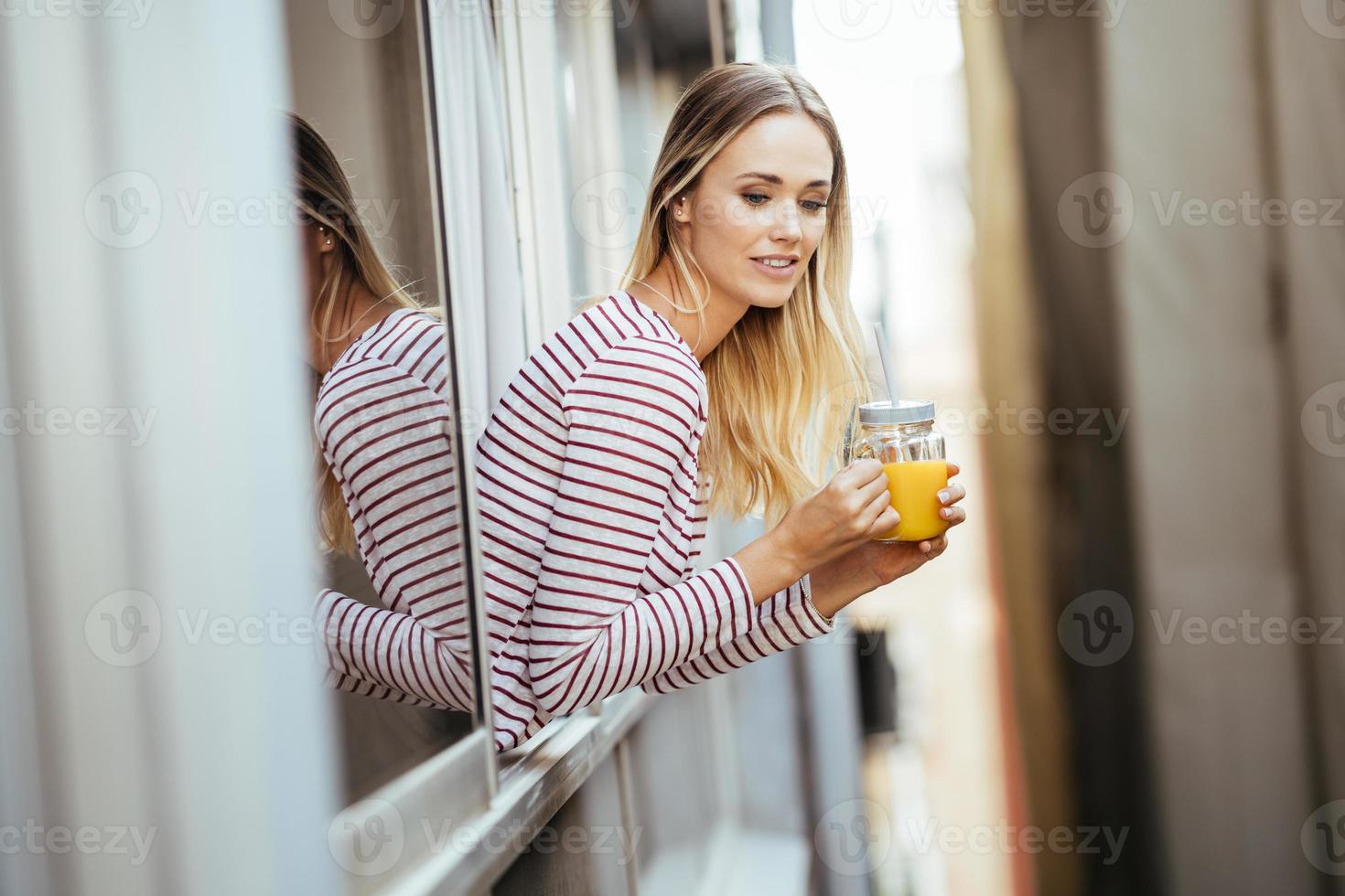 ung kvinna dricker ett glas naturlig apelsinjuice, lutad ut genom fönstret i hennes hem. foto