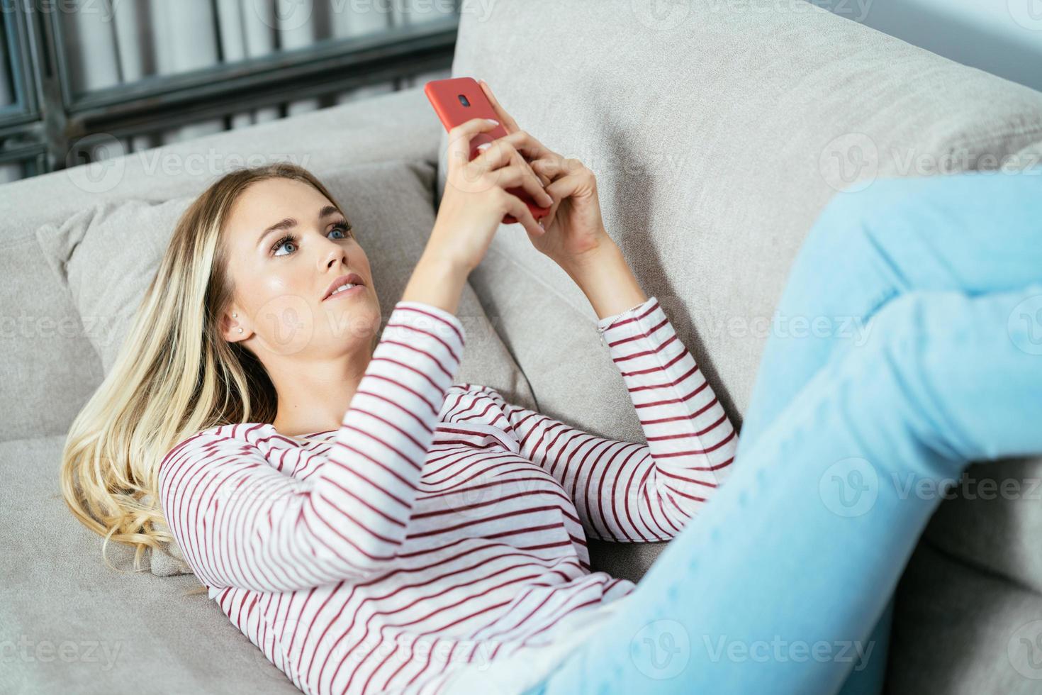 ung kaukasisk kvinna med sin smartphone liggande på soffan. foto