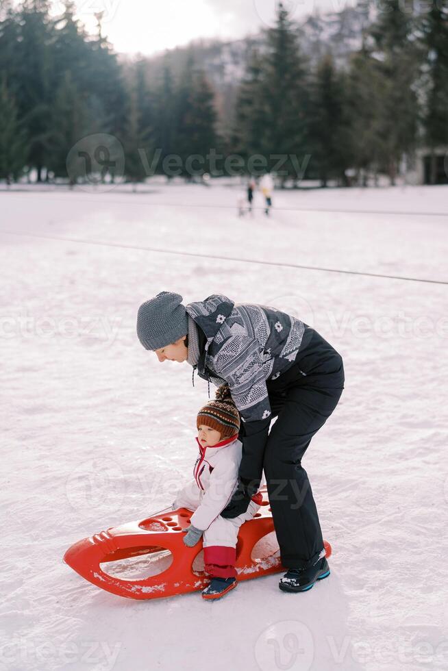 mamma sätter en liten flicka på en kälke på en snöig gräsmatta foto