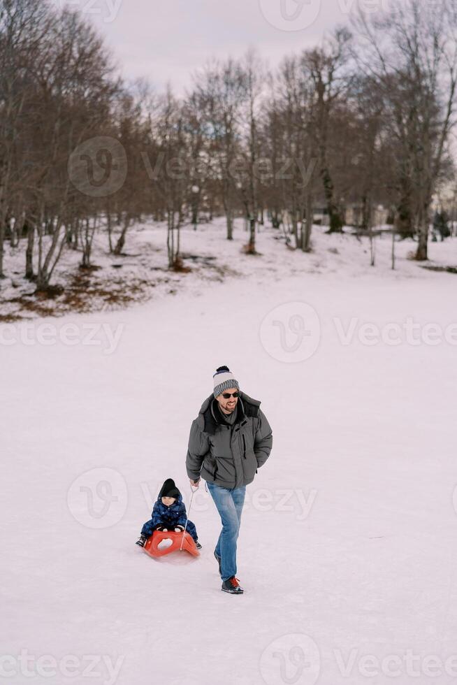 pappa bär en liten pojke på en släde tvärs över en snöig enkel nära de skog foto