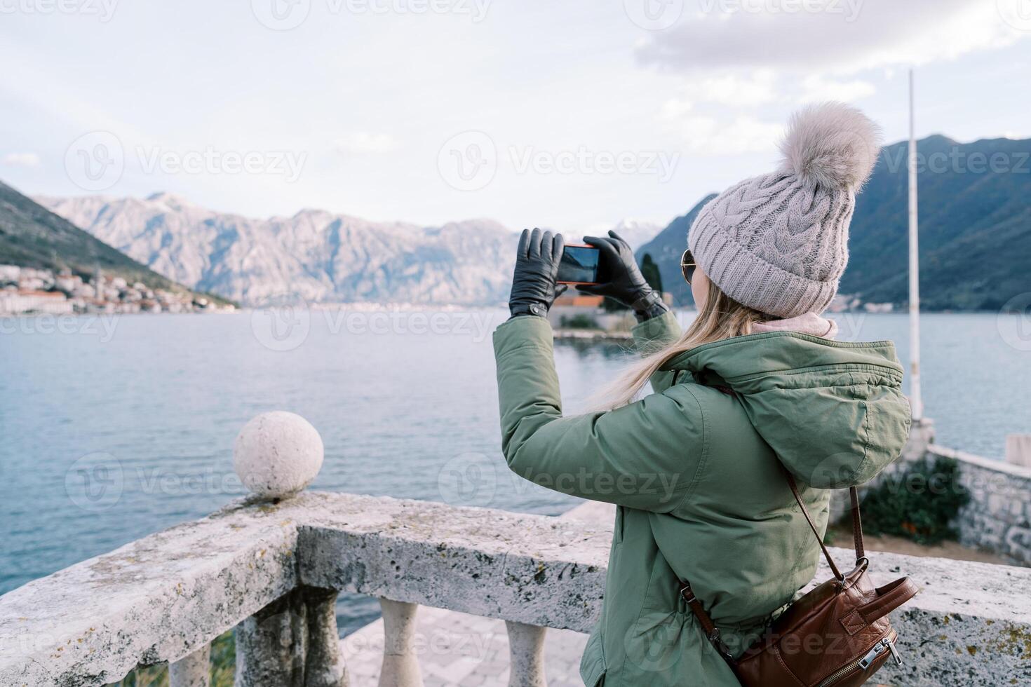 flicka med en ryggsäck skjuter de hav och en berg räckvidd med en smartphone medan stående på en sten balustrad. sida se foto