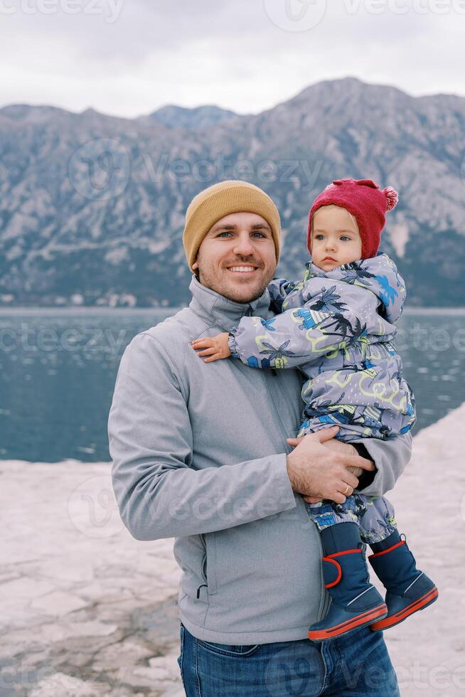 leende pappa med en liten flicka i hans vapen står på de havsstrand mot de bakgrund av bergen foto