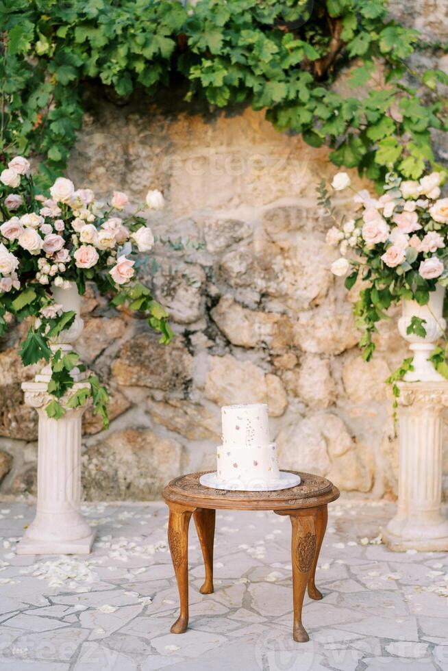 bröllop kaka står på en trä- tabell nära en bröllop halvbåge nära en sten vägg i de trädgård foto