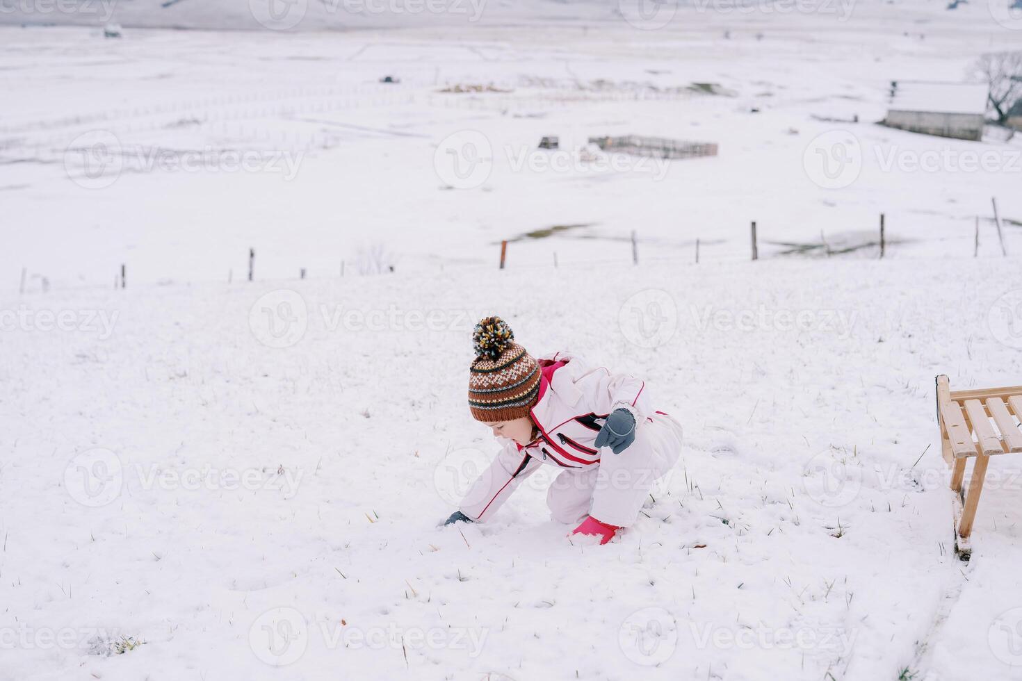 liten flicka gör en snöboll medan hukande på en snöig clearing nära en trä- släde foto
