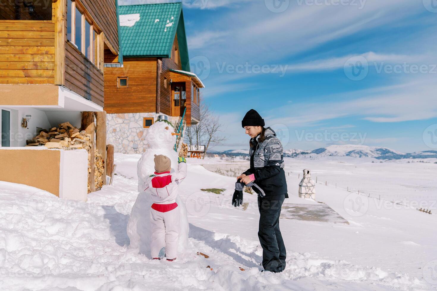liten flicka dekorerar en snögubbe nära de stuga medan stående Nästa till henne mor sätta på vantar foto