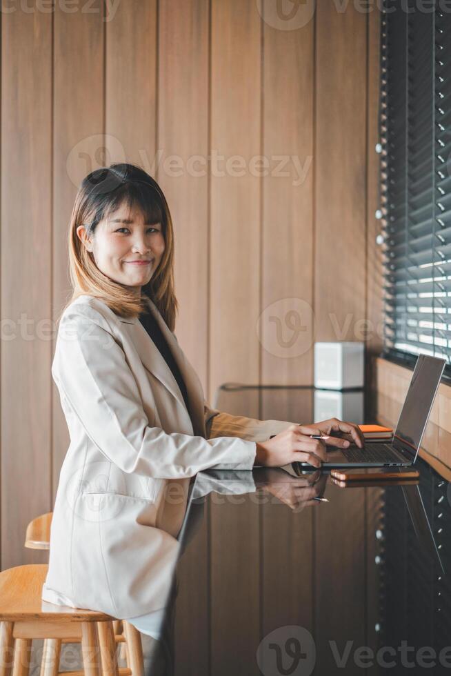 en självsäker ung professionell Arbetar på henne bärbar dator förbi en fönster med persienner, henne redo uppträdande reflekterande effektivitet och ambition i en företags- miljö. foto