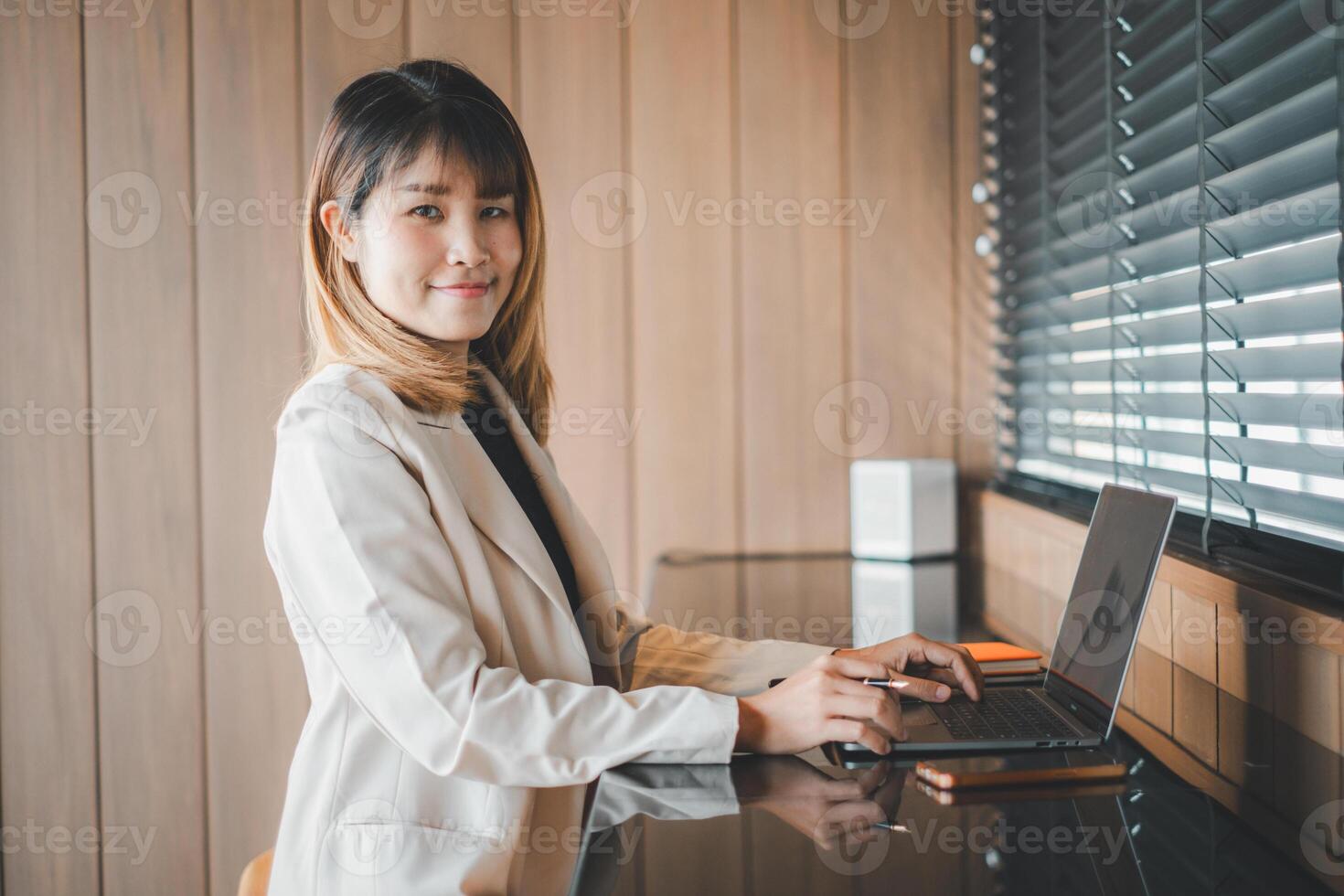en självsäker ung professionell Arbetar på henne bärbar dator förbi en fönster med persienner, henne redo uppträdande reflekterande effektivitet och ambition i en företags- miljö. foto