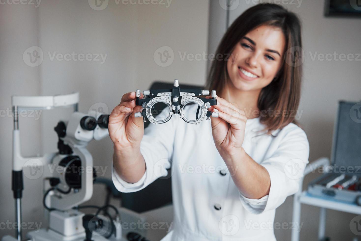 annan utrustning i rummet. ung attraktiv kvinnlig ögonläkare med speciell anordning för att testa ögon som står på kontoret foto