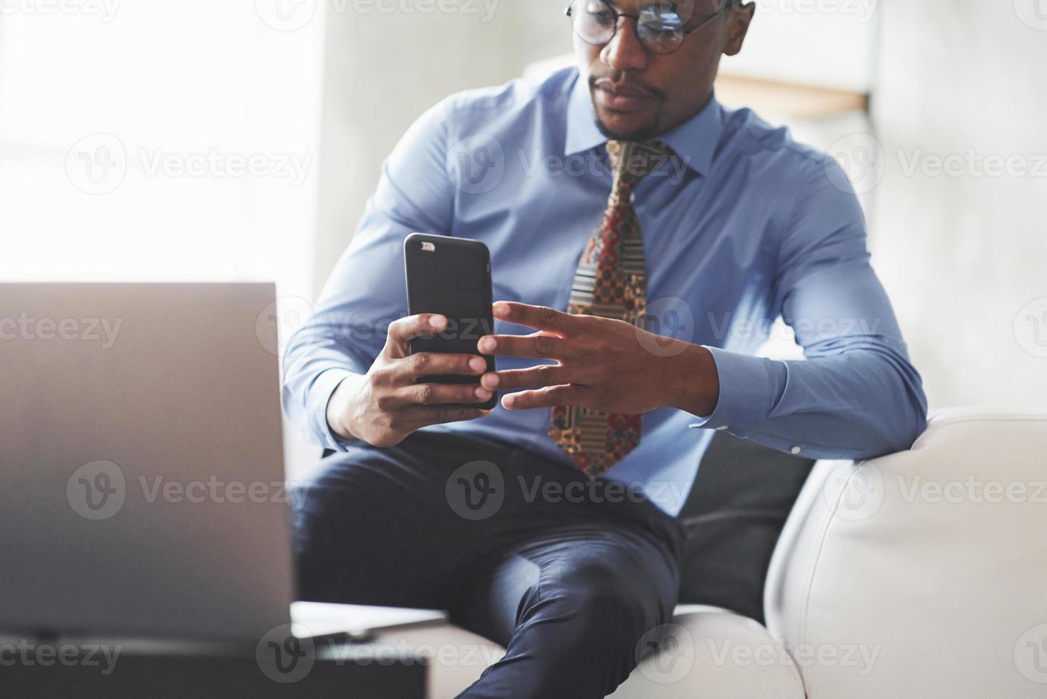 paustid. foto av ung snygg svart man i kostym och glasögon som håller och tittar på telefonen medan han sitter i soffan