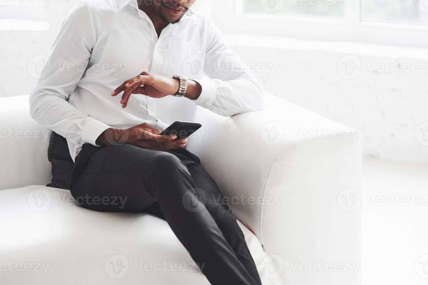 beskuret foto. kontrollerar tiden. ung afroamerikansk kille i kontorskläder sitter på den vita soffan foto