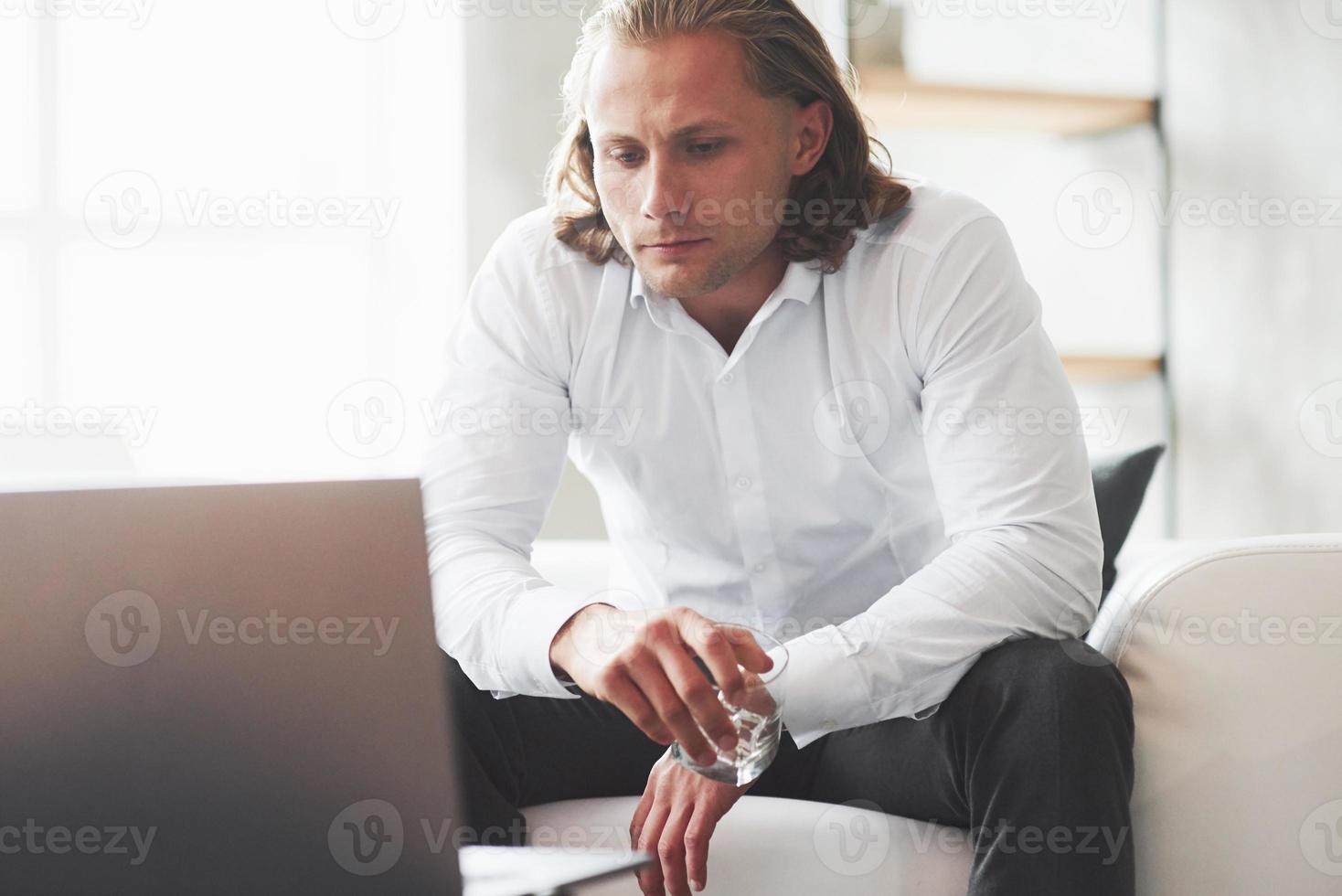 ser hur processen går. allvarlig ung affärsman sitter inomhus i soffan med whiskyglas foto