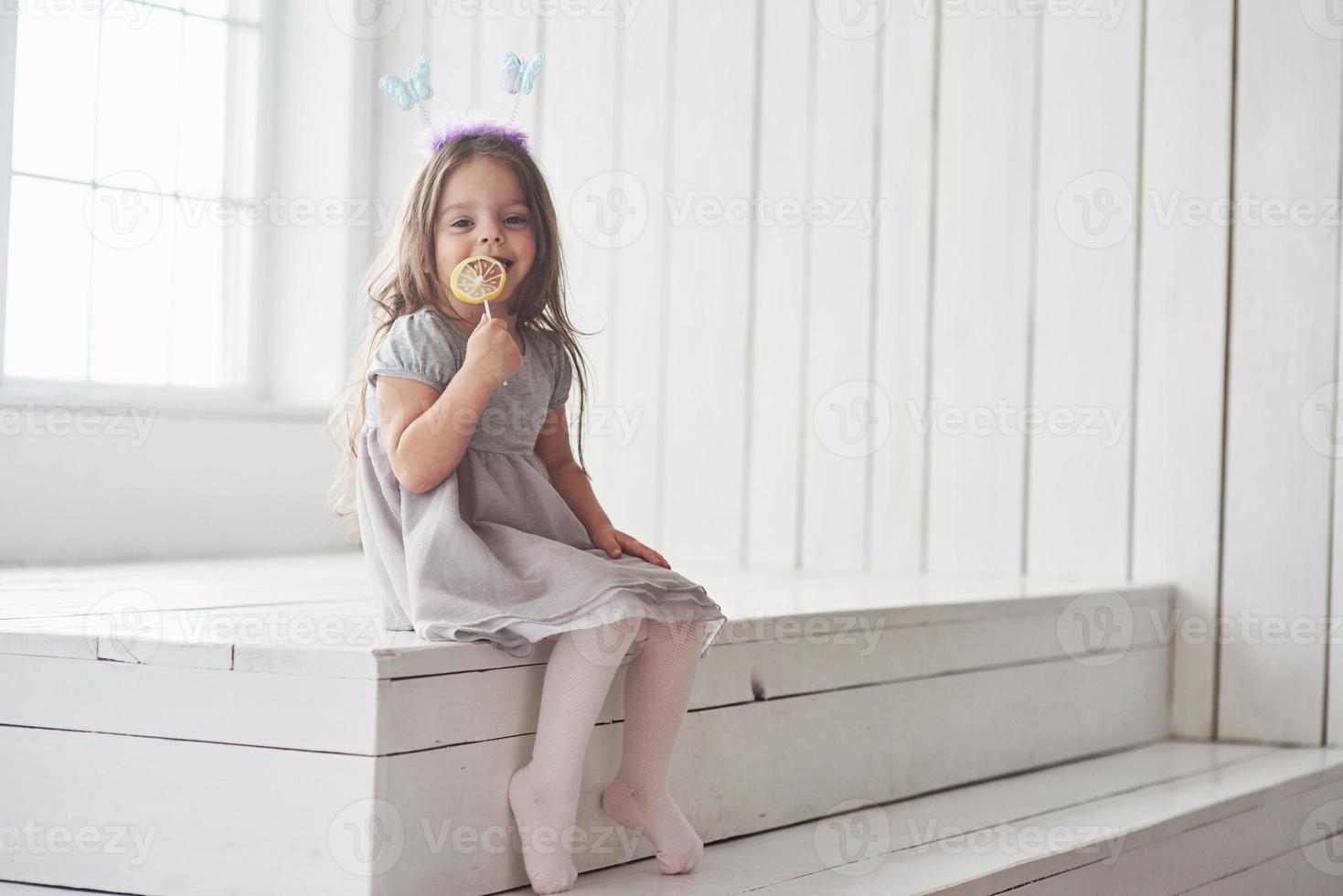 sitter på trappan. söt liten flicka som äter det gula ljuset med leksaksfjärilar på huvudet i det vita rummet foto