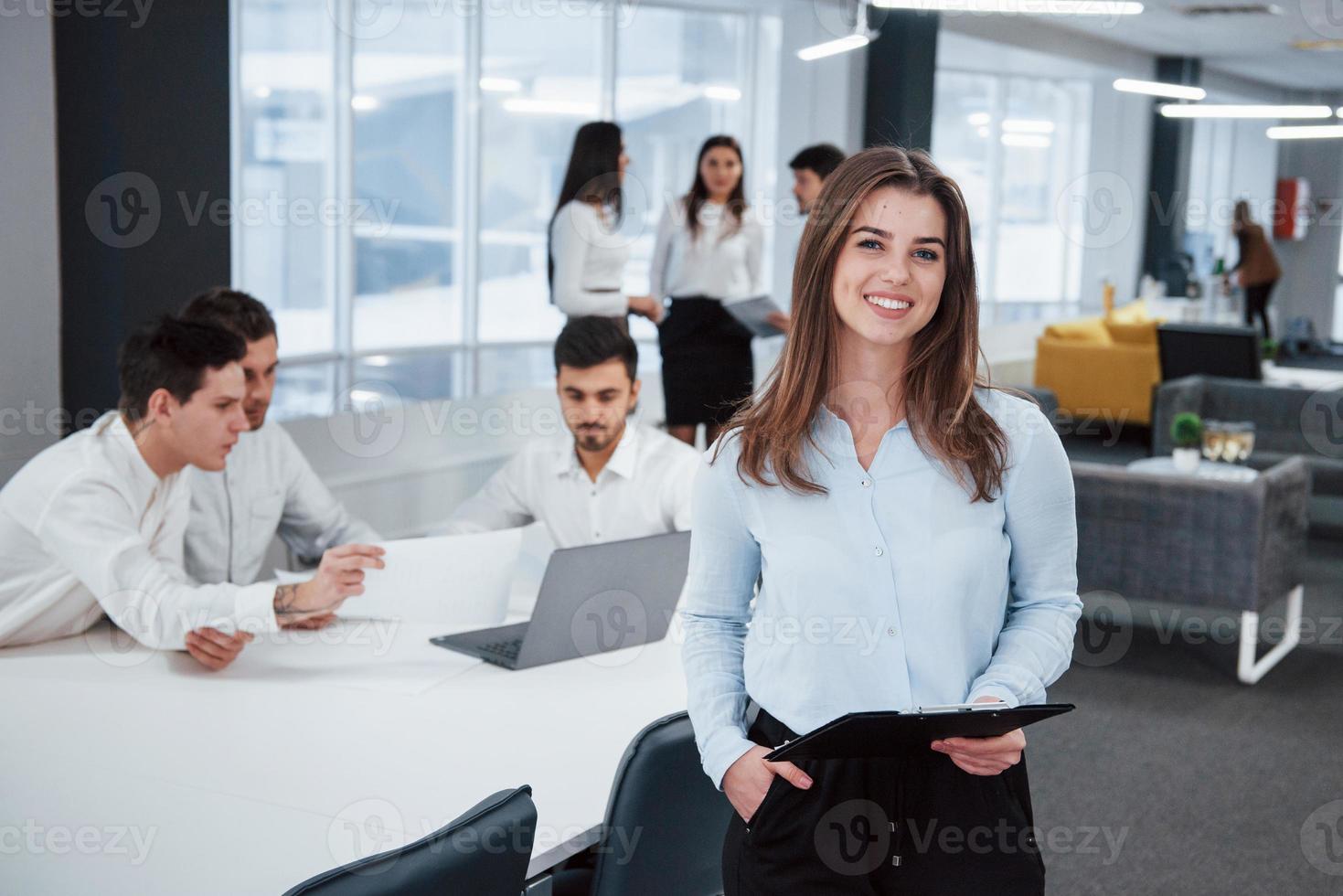 avslappnad kvinna inomhus. porträtt av ung flicka står på kontoret med anställda i bakgrunden foto