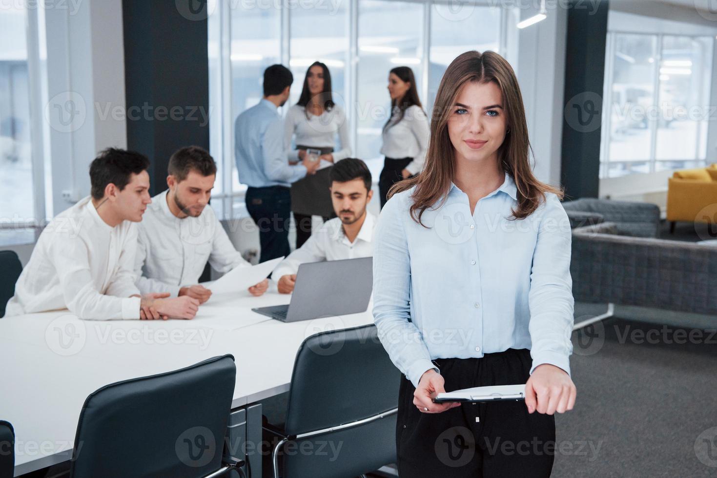 titta rakt in i kameran. porträtt av ung flicka står på kontoret med anställda i bakgrunden foto