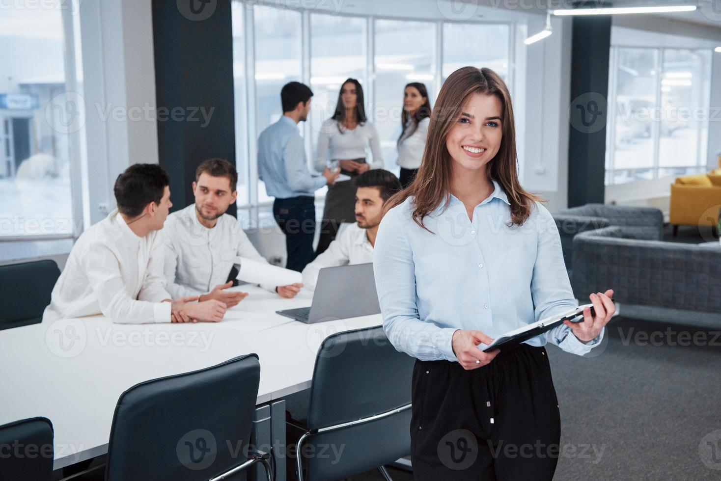 mår bra för hon älskar sitt jobb. porträtt av ung flicka står på kontoret med anställda i bakgrunden foto
