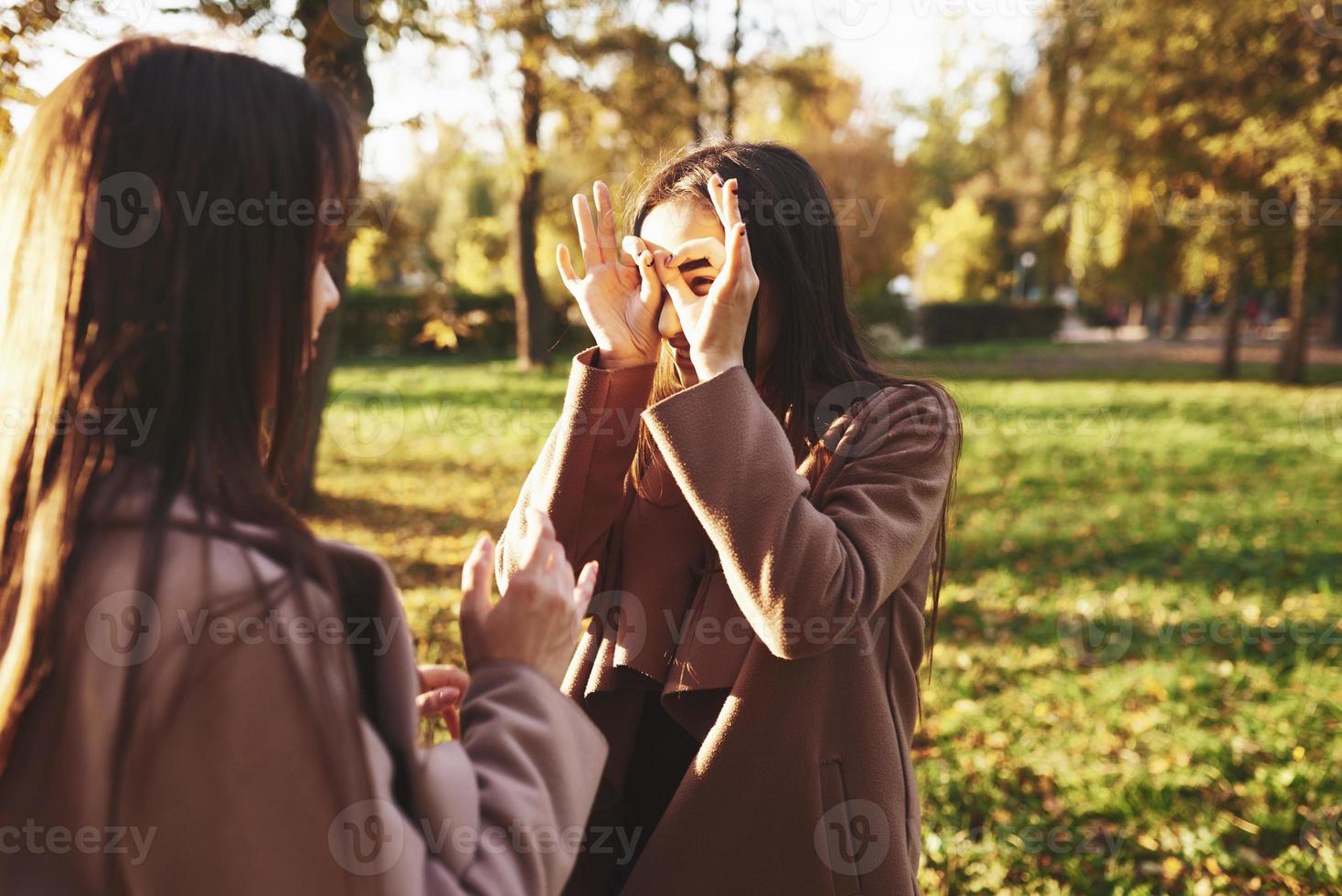 två brunett tvillingsystrar som står framför varandra och har roligt och gör cirklar med fingrar runt ögonen, bär avslappnad kappa i solig höstpark på suddig bakgrund foto