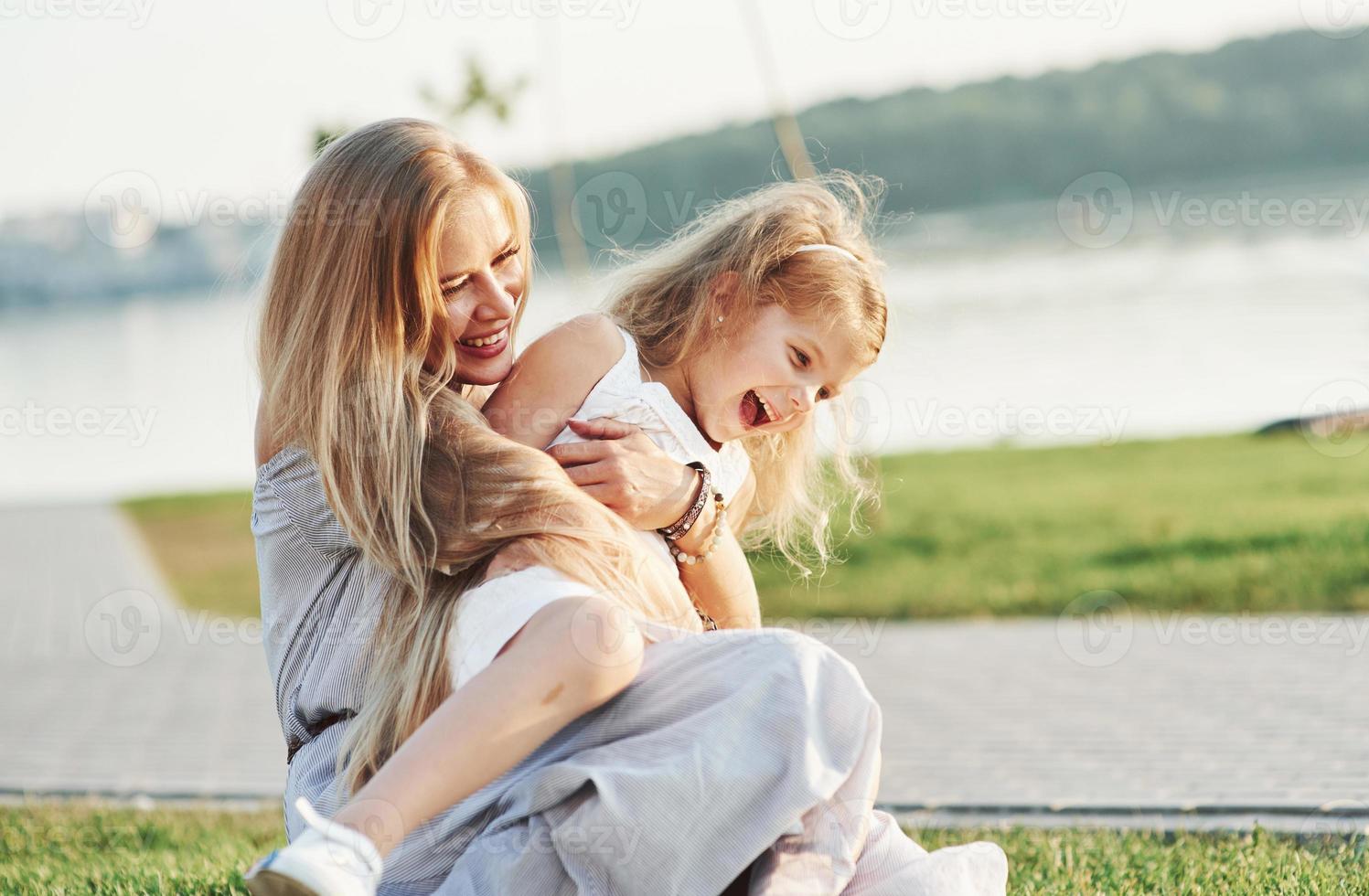 ren kärlek. foto av ung mor och hennes dotter har bra tid på det gröna gräset med sjön i bakgrunden