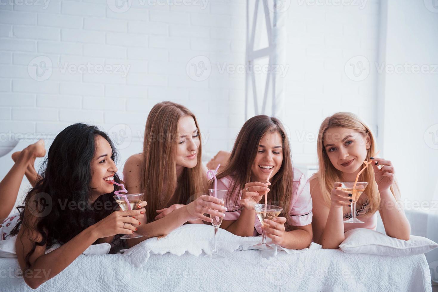 håller glasögon med alkohol. glada flickor i nattkläder ligger på sängen i vita rum och har firande foto