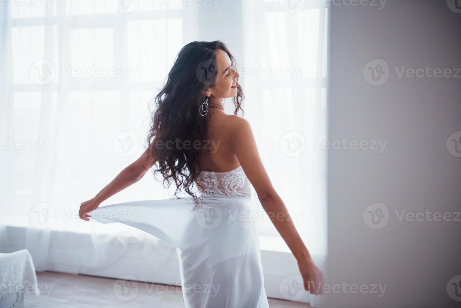bakifrån. vacker kvinna i vit klänning står i vitt rum med dagsljus genom fönstren foto