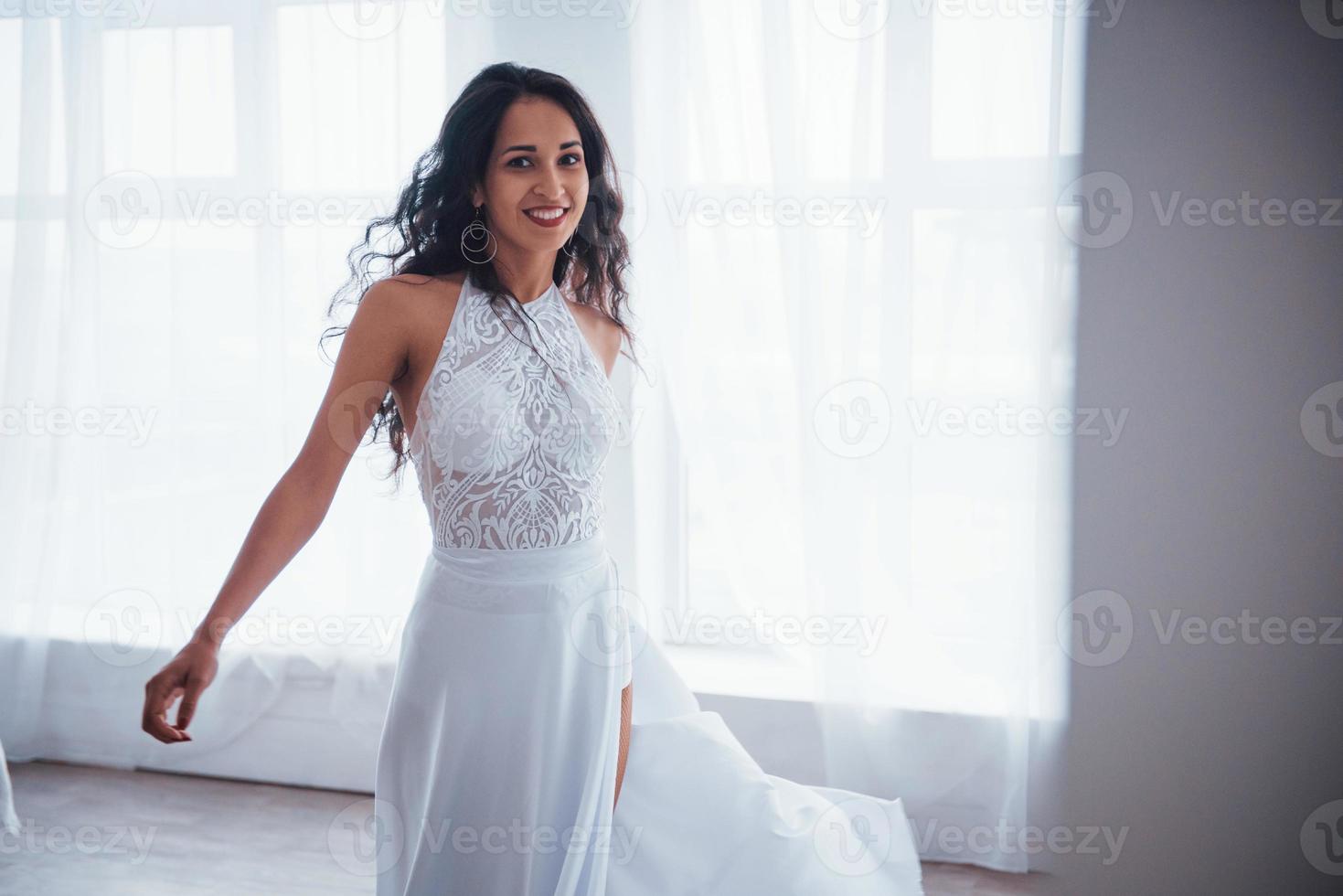 attraktiv tjej. vacker kvinna i vit klänning står i vitt rum med dagsljus genom fönstren foto