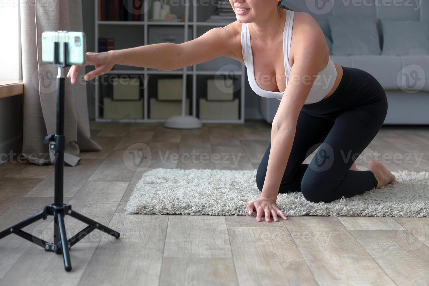 kvinna Sammanträde på de golv i yoga byxor inspelning ett uppkopplad kondition med henne telefon och stativ, leende på de kamera i en levande rum miljö foto