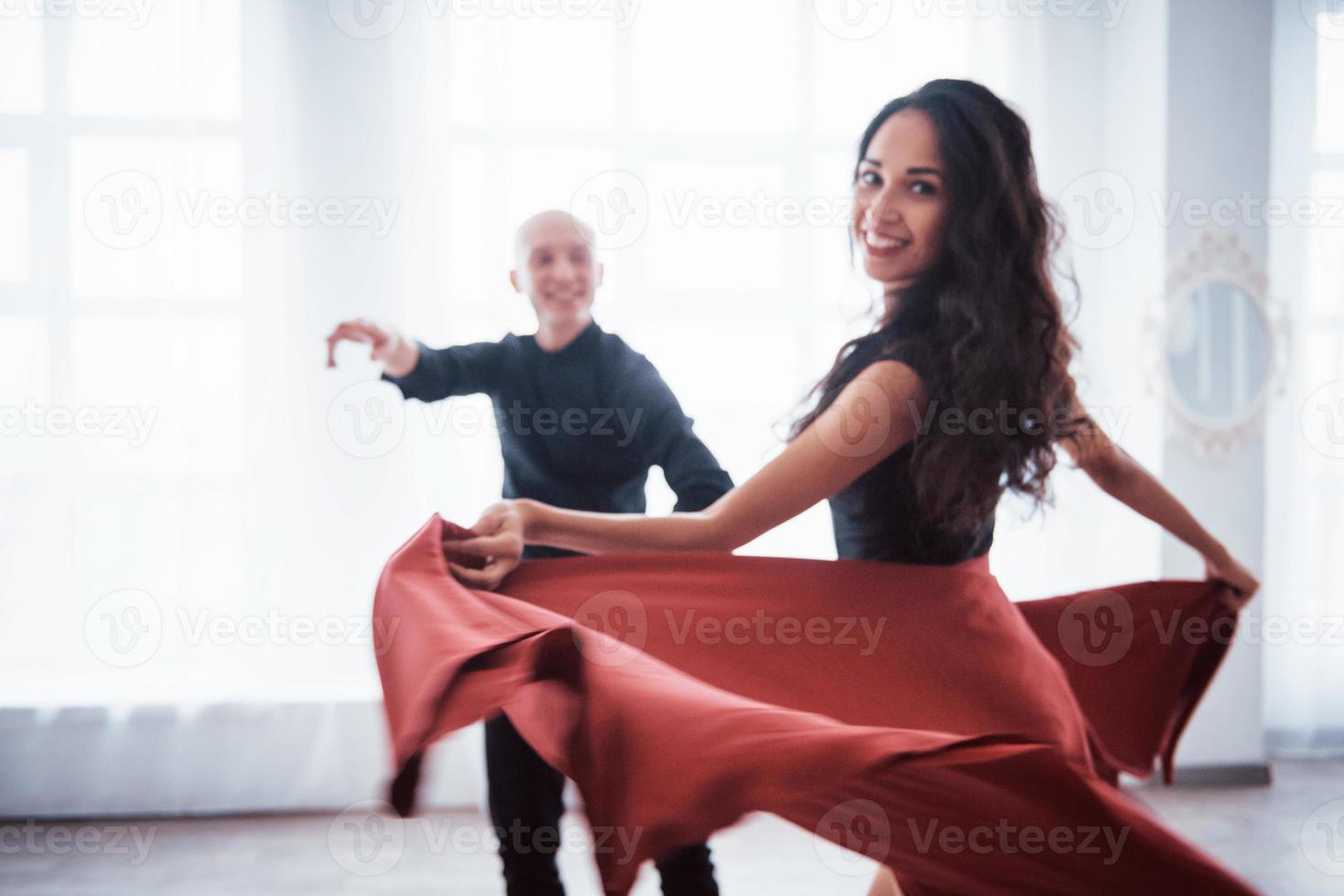 njuter av att göra föreställningen. ung vacker kvinna i röda och svarta kläder dansar med skallig kille i det vita rummet foto