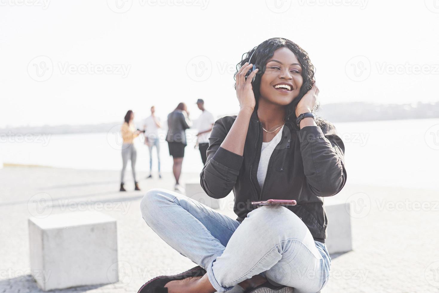 porträtt av en vacker ung söt afroamerikansk flicka som sitter på stranden eller sjön och lyssnar på musik i sina hörlurar foto