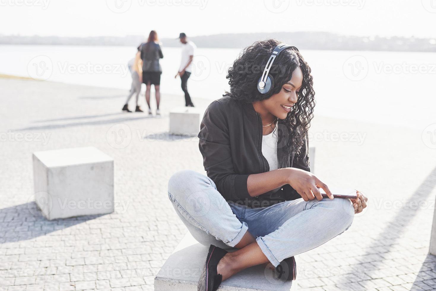 porträtt av en vacker ung söt afroamerikansk flicka som sitter på stranden eller sjön och lyssnar på musik i sina hörlurar foto