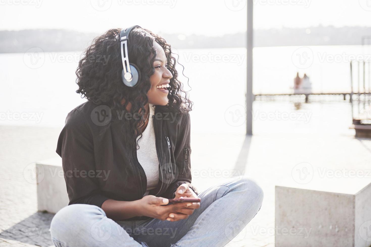 porträtt av en vacker ung söt afroamerikansk flicka som sitter på stranden eller sjön och lyssnar på musik i sina hörlurar foto