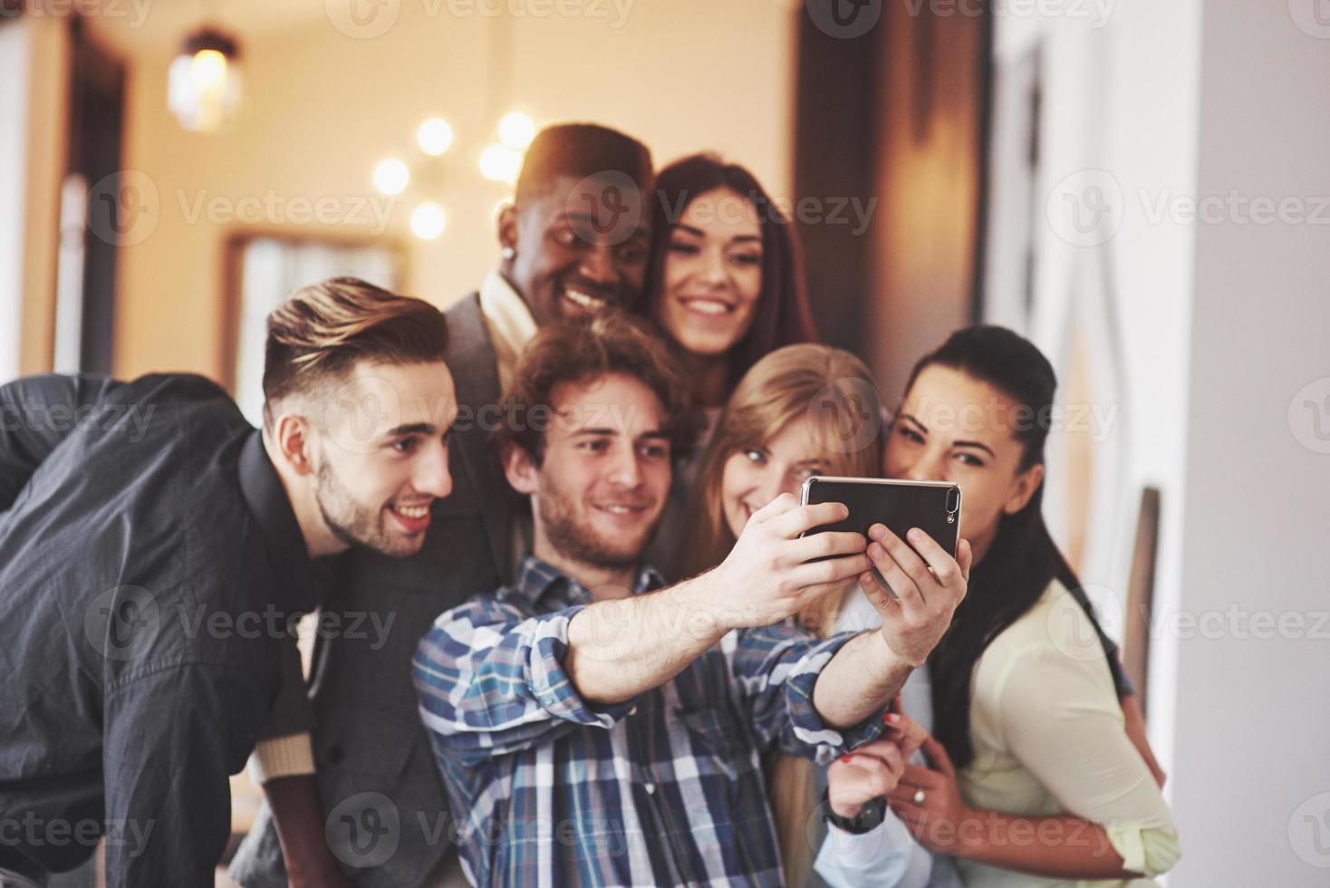 vänner har kul på restaurang. tre pojkar och tre tjejer gör selfie och skrattar. på förgrunden pojke håller smart telefon. alla bär vardagskläder foto