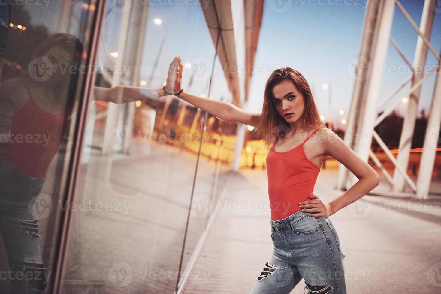 vacker sexig turist flicka går i livlig stadsgata nära flygplatsen. kvinna tittar på kameran utomhus klädd i moderiktig röd t-shirt foto